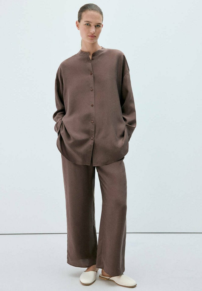 Personne debout portant une chemise marron ample boutonnée et un pantalon large assorti avec des chaussures blanches slip-on sur fond blanc.