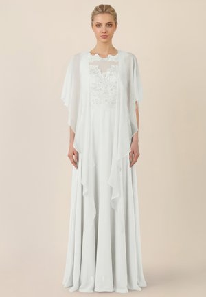 Vrouw die een vloeiende witte jurk draagt met kanten details op de borst en doorschijnende, gedrapeerde mouwen, staand tegen een neutrale achtergrond.