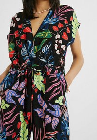 Blommig jumpsuit med korta ärmar, som har livfulla gröna, röda och blå blad på en svart bakgrund. Elastisk midja och fickor ingår.