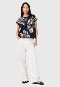 Camiseta negra de flores con mangas cortas y un corte holgado, combinada con pantalones anchos blancos y sandalias negras, frente a un fondo neutro.
