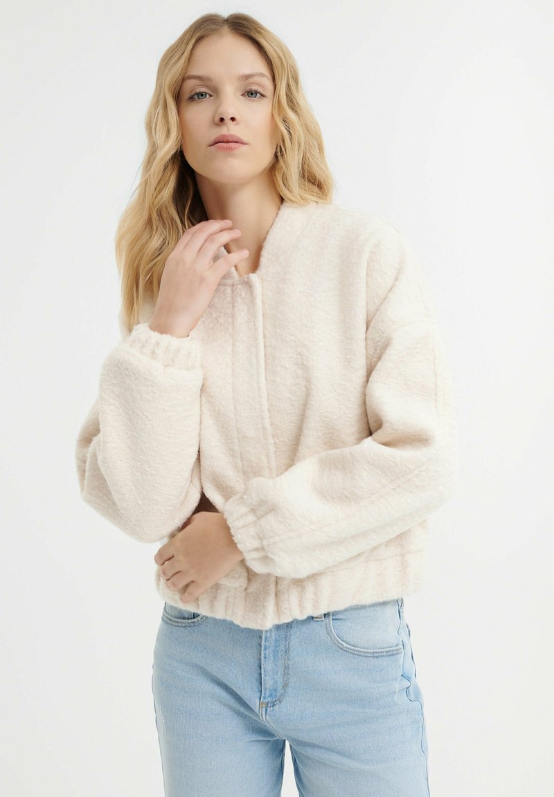 Przycięta, puszysta kurtka w jasnym beżu z prążkowanymi mankietami i dołem; ma okrągły dekolt oraz zapięcie z przodu bez zamków błyskawicznych ani guzików.