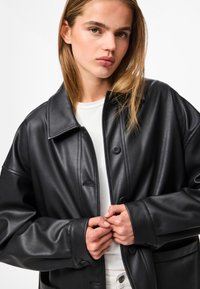 Schwarze Lederjacke mit glatter Textur, spitzen Kragen und Knopfverschluss, über einem weißen Hemd getragen.