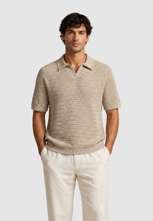 Uomo in piedi con le mani nelle tasche, indossa una polo lavorata a maglia beige a maniche corte e pantaloni chiari su sfondo semplice.