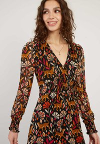 Vrouw in een zwart langemouwen jurk met oranje tijger- en bloemenpatronen, glimlachend en naar de zijkant kijkend tegen een effen achtergrond.