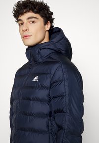 Mørkeblå puffjakke med lynlås foran, hætte og horisontale quiltede sektioner. Har en hvid Adidas-logo på venstre bryst.