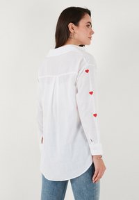 White long-sleeve shirt with red heart accents on the sleeves, lightweight fabric, button cuffs, and a curved hem. Suitable for casual wear.