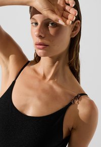 Maillot de bain noir avec des bretelles fines, un encolure dégagée et une finition texturée. Détail de matériel remarquable sur la bretelle relié par un fermoir.