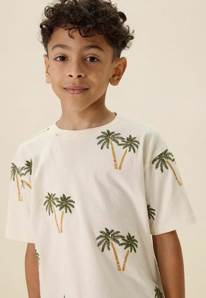 Jongen met krullend haar, gekleed in een wit T-shirt met groene en gele palmboompatronen, staand tegen een effen beige achtergrond.
