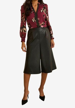 Woman wearing a long-sleeve maroon patterned blouse with black studded trim, black wide-leg culottes, and black pointed high heels.