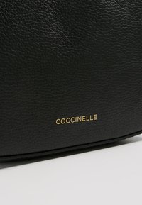 Černá kožená kabelka s texturovaným povrchem a zlatým logem "Coccinelle". Má elegantní, zaoblený tvar a minimalistický design.