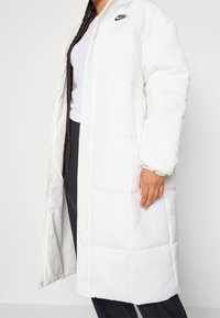 Vadderad vit lång kappa med dragkedja fram, ståkrage och svart Nike-logotyp. Har elastiska ärmslut och två sidofickor.