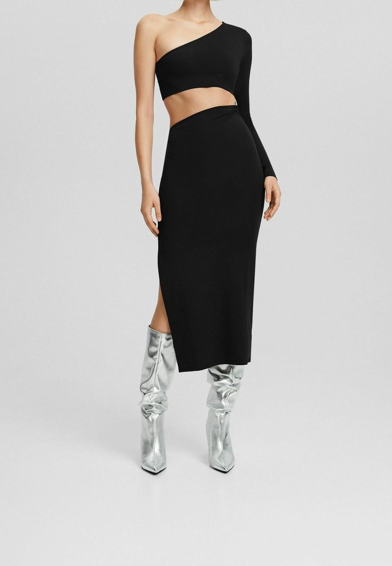 Schwarzes zweiteiliges Outfit mit einem asymmetrischen One-Shoulder-Oberteil und einem Midirock mit seitlichem Schlitz. Kombiniert mit metallisch silbernen kniehohen Stiefeln.