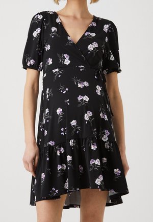 Femme portant une robe portefeuille noire à fleurs avec des manches courtes bouffantes et un ourlet à volants, ornée de fleurs violet clair et blanches.