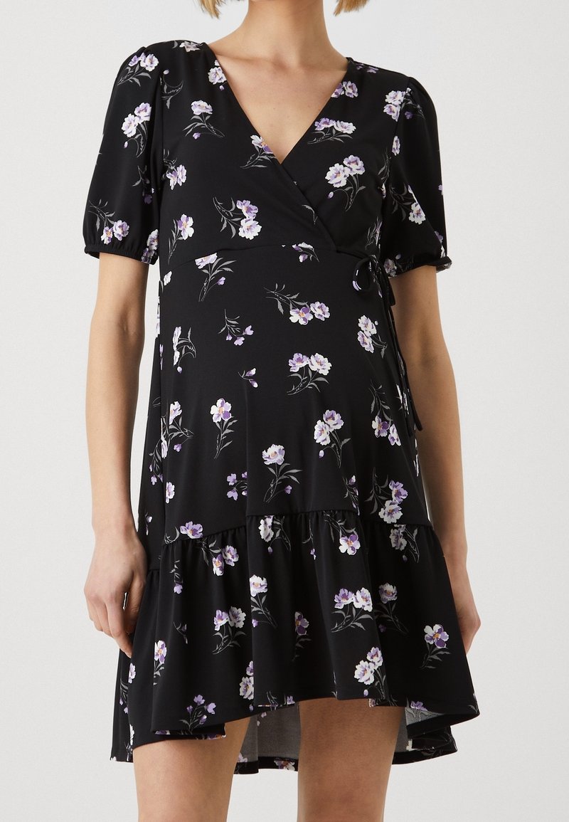 Femme portant une robe portefeuille noire à fleurs avec des manches courtes bouffantes et un ourlet à volants, ornée de fleurs violet clair et blanches.