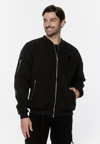 Czarna bomberka z zamkiem z przodu, ściągaczami przy mankietach oraz dwoma zasuwanymi kieszeniami po bokach. Posiada mały, okrągły emblemat na piersi.