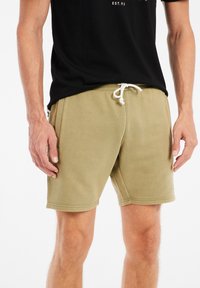 Khaki zwemschoenen gemaakt van zacht materiaal, met een elastische tailleband en witte trekkoord, zijzakken en een minimalistisch ontwerp.