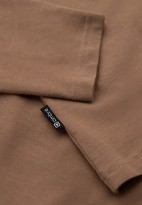 Braunes Baumwoll-T-Shirt-Gewebe mit kurzen Ärmeln, das sichtbare Nähte und ein schwarzes Etikett mit weißem Text aufweist.