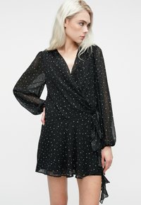 Kvinde med langt blondt hår iført en sort gennemsigtig kjole med langærmer og hvide stjerne mønstre, poserende med den ene hånd på hoften og ser til højre.