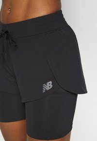 Närbild på en person som bär svarta lager av träningsshorts med dragsko i midjan och en reflekterande New Balance-logotyp p�å det yttre lagret.