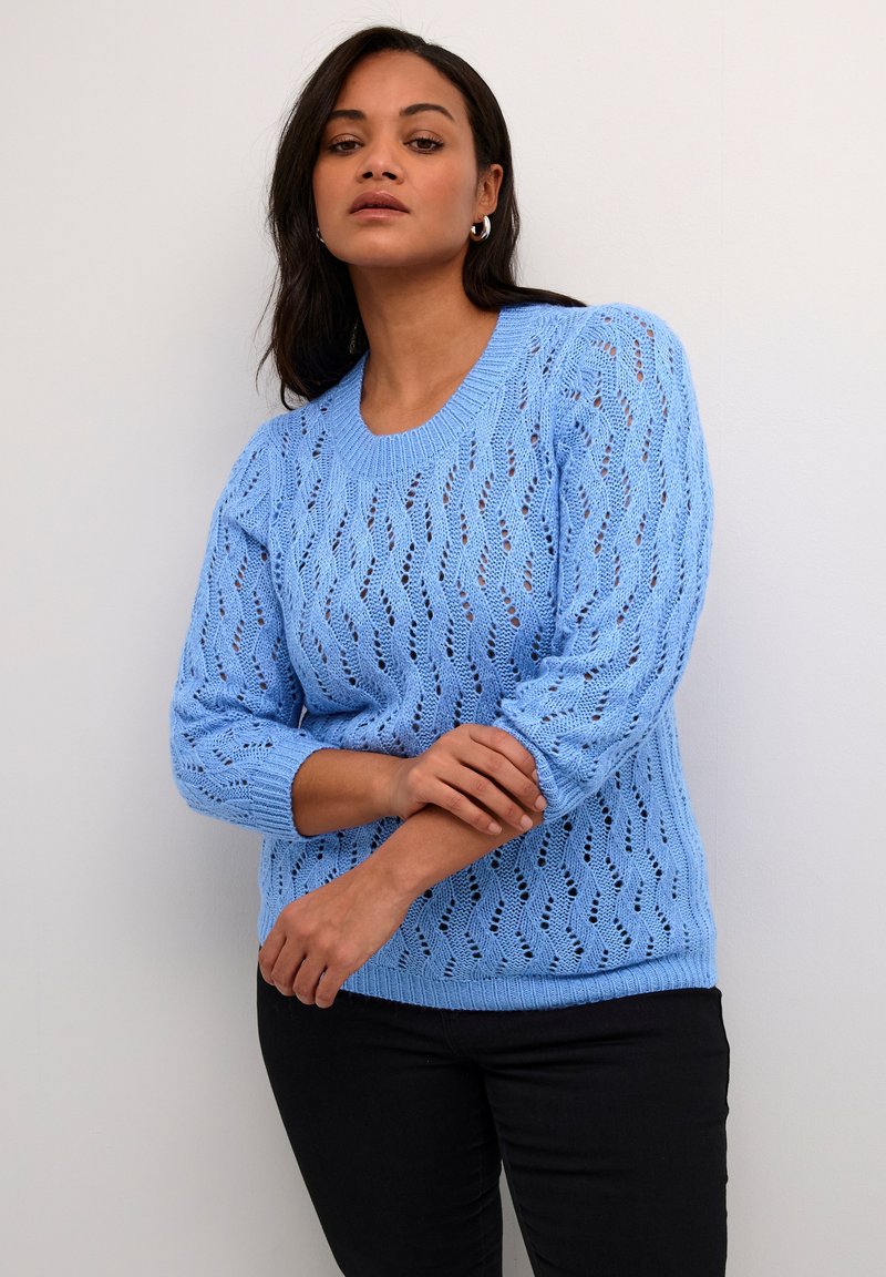 Hellblauer gestrickter Pullover mit einem strukturierten, offenen Design, ausgestattet mit einem runden Ausschnitt und Dreiviertelärmeln, kombiniert mit dunklen Hosen.