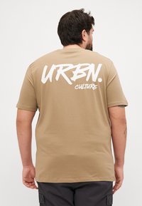 Korte ermer tan t-skjorte med hvit trykt tekst "URBN. CULTURE" på ryggen. Laget av myk bomull med løs passform.