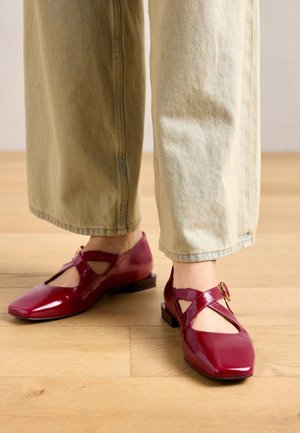 Persoon die beige broek en glanzende bordeauxrode Mary Jane-schoenen met gespbanden draagt, staand op een houten vloer.