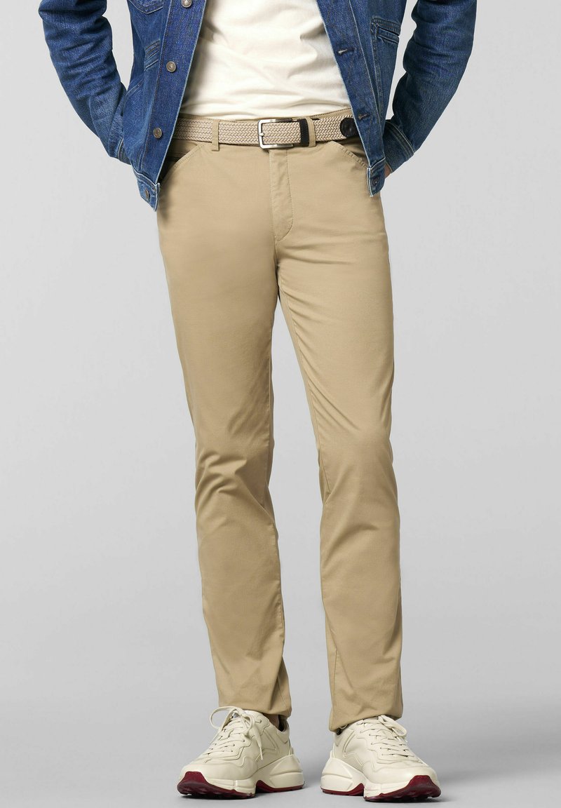 Beige Chinos mit schmaler Passform, strukturiertem Stoff, Seitentaschen und einem geflochtenen Gürtel. Kombiniert mit weißen Sneakers mit roten Akzenten.