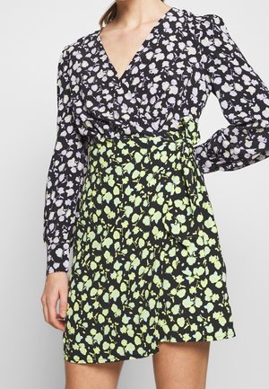 Frau trägt ein Wickelkleid mit schwarzem Hintergrund, lila Blumenoberteil und grünem Blumensrock, posiert mit einer Hand in der Hüfte.