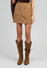 Jupe marron avec deux poches avant et des boutons décoratifs. Portée avec des bottes de cowboy en daim marron avec un haut replié et un bout pointu.