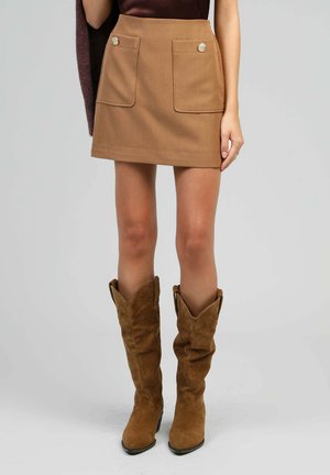 Jupe marron avec deux poches avant et des boutons décoratifs. Portée avec des bottes de cowboy en daim marron avec un haut replié et un bout pointu.