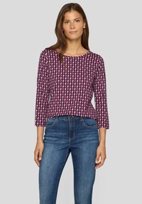 Frau in einem langärmeligen Oberteil mit einem geometrischen Muster in Pink, Lila und Schwarz, kombiniert mit blauen Jeans, steht vor einem einfarbigen hellgrauen Hintergrund.