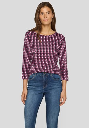 Frau in einem langärmeligen Oberteil mit einem geometrischen Muster in Pink, Lila und Schwarz, kombiniert mit blauen Jeans, steht vor einem einfarbigen hellgrauen Hintergrund.