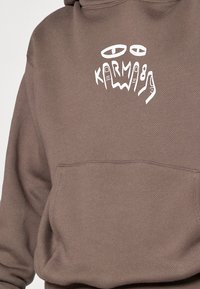 Hoodie marron avec une poche frontale, présentant un design graphique blanc comprenant des yeux et le texte "KARMA89." Le texte est stylisé et ludique.