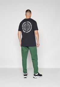 Sort t-shirt med cirkulært hvidt "Under Armour" logo på ryggen, parret med grønne sweatpants og sorte træningssko.
