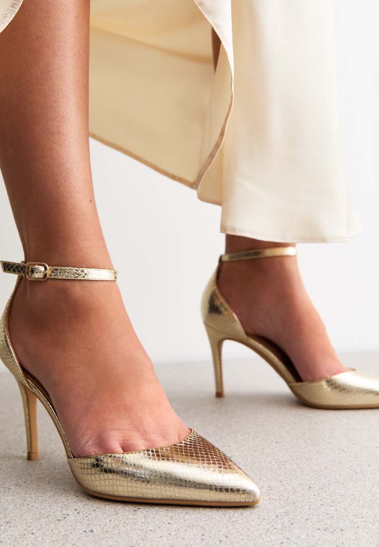 Scarpe dorate con tacco alto, punta affusolata e cinturino alla caviglia; superficie texturizzata simile a quella di un rettile; tacco sottile; indossate con una gonna di colore chiaro.