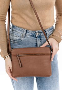 Borsa a tracolla in pelle marrone con una texture liscia, tasca anteriore con zip e tracolla regolabile, indossata con jeans azzurri chiari.