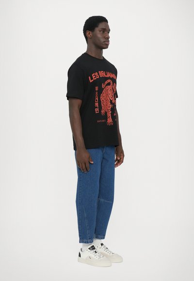 Black cotton t-shirt with red graphic print featuring a lion. Paired with blue denim trousers and white sneakers. Model stands in profile.