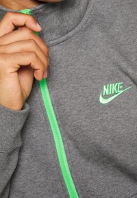 Šedá mikina na zip s jasně zeleným zipem. Na hrudi se nachází malé zelené logo Nike. Materiál vypadá měkce a je texturovaný.