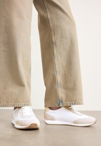 Baskets blanches avec une accent en daim beige, fermeture à lacets plates et semelle en caoutchouc, associées à un jean droit beige clair.