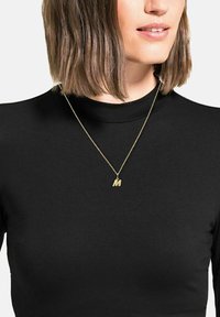 Collana d'oro con un ciondolo a forma di lettera "M", indossata sopra una maglietta nera a collo alto. La catena è sottile, con un design semplice.
