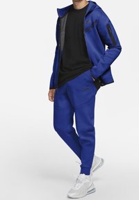 Niebieski dres Nike z zippowaną kurtką z kapturem i czarnymi akcentami, w połączeniu z dopasowanymi spodniami; noszony z białymi butami sportowymi.