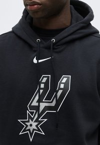 Άνδρας που φοράει μαύρο φούτερ με κουκούλα με το λογότυπο της Nike και το έμβλημα της ομάδας καλαθοσφαίρισης San Antonio Spurs στο μπροστινό μέρος.