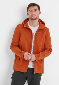Orange softshell jacket with a hood, featuring zipper pockets, flat seams, and a logo on the chest. Worn over a white t-shirt.