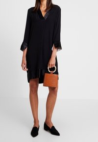 Svart klänning med V-ringning och trekvartslånga ärmar, prydd med satin detaljer vid halsringningen och fållen. Håller en strukturerad handväska i orange.
