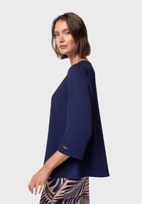 Blusa blu navy con maniche a tre quarti, texture liscia, scollatura rotonda, con una sottile tasca laterale e dettagli in metallo argentato.
