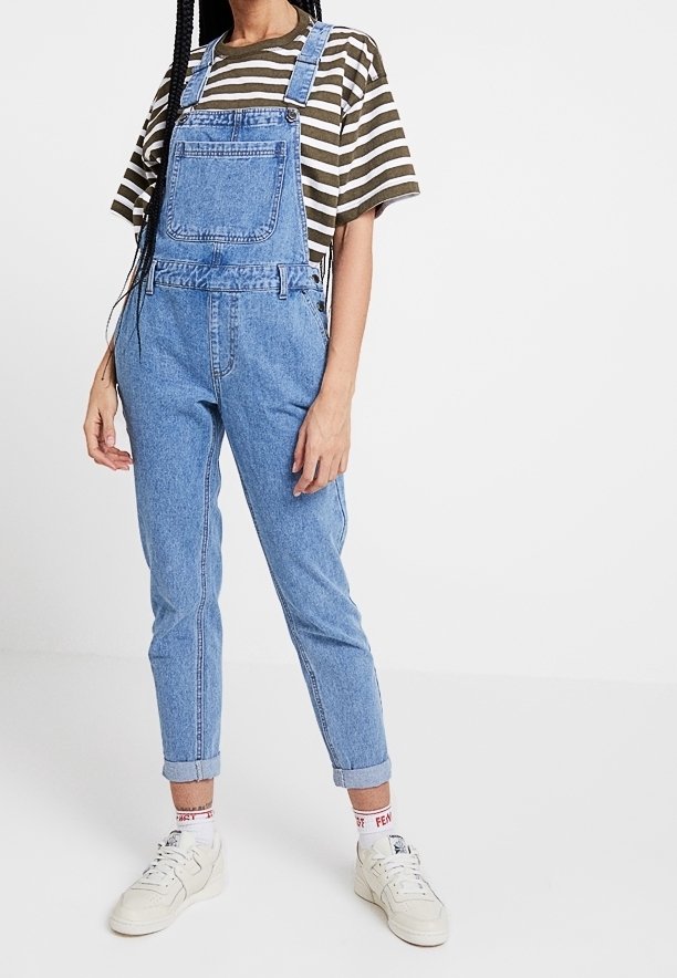 Salopette en denim bleu avec bretelles ajustables et poche avant, assortie d'un t-shirt rayé vert et blanc. Des baskets blanches complètent le look.