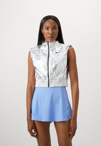 Silberne ärmellose Jacke mit hohem Kragen und Reißverschluss, kombiniert mit einem hellblauen, geflechteten Rock. Strukturstoff und sportliches Design.