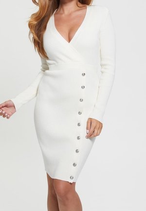 Mujer que lleva un vestido cruzado de punto acanalado blanco con mangas largas y una línea de botones plateados en el lateral.
