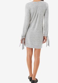 Vestido de suéter gris de punto con mangas largas, acentos de lazo en los puños y un pliegue en la parte posterior. Presenta un corte recto y una textura suave.
