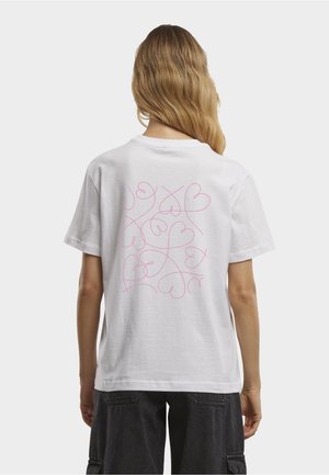 Person med langt blondt hår som har på seg en hvit T-skjorte med rosa abstrakt hjerte-linjearbeid på ryggen, kombinert med svarte cargoshorts.